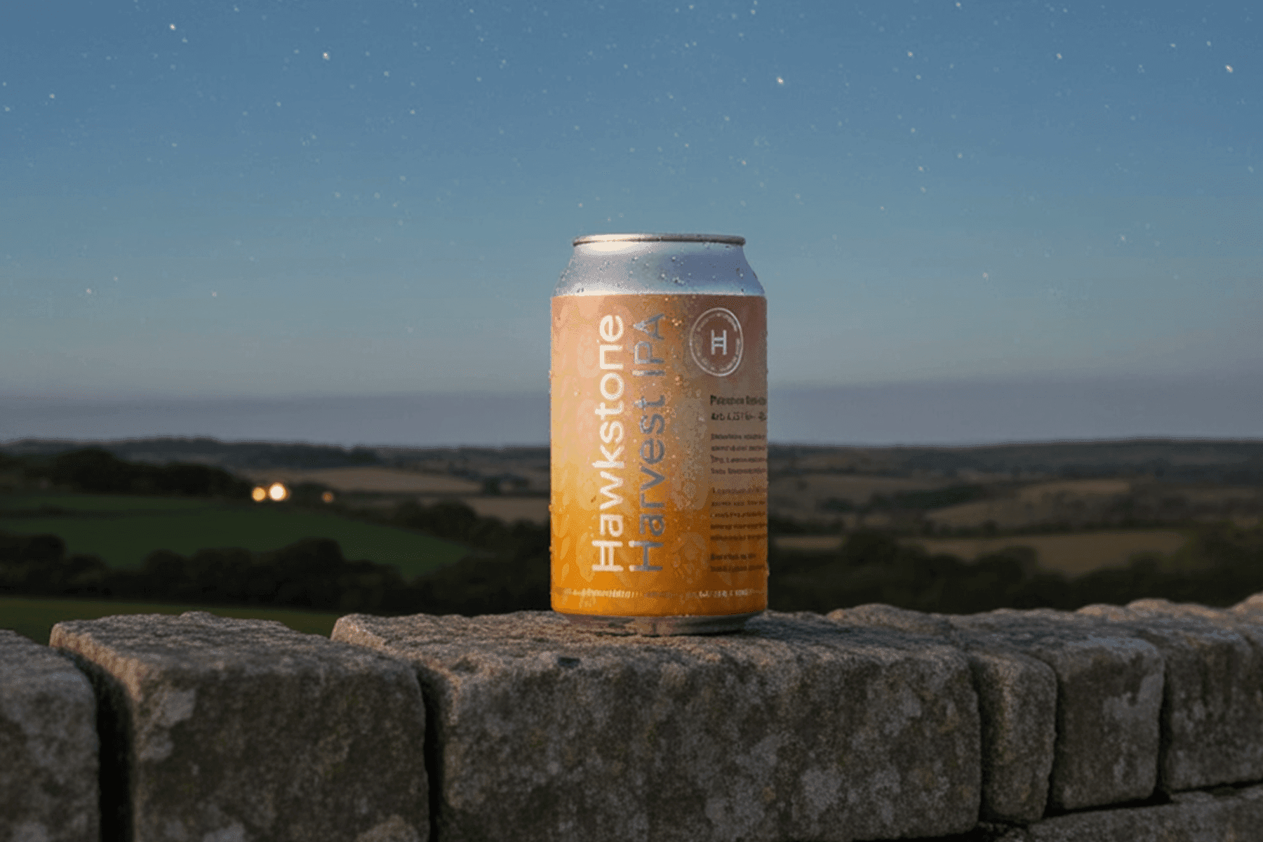 A can of Hawkstone Harvest IPA beer, covered in condensation, rests on a rustic stone wall at twilight. The can is silver at the top and bottom, with an orange label displaying "Hawkstone Harvest IPA" in white text. In the background, a serene landscape of rolling hills and fields extends under a starry, dark blue sky, with faint lights visible in the distance